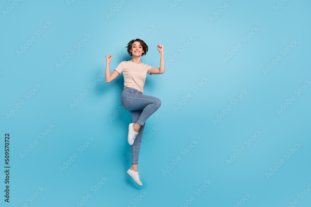 Full length photo of pretty lucky woman wear beige t-shirt jumping high rising fists empty space isolated blue color background