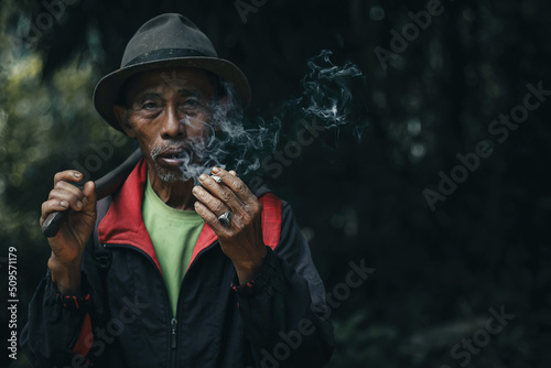 Close up of Asian farmer elderly man is smoking while carrying hoe after work on field 