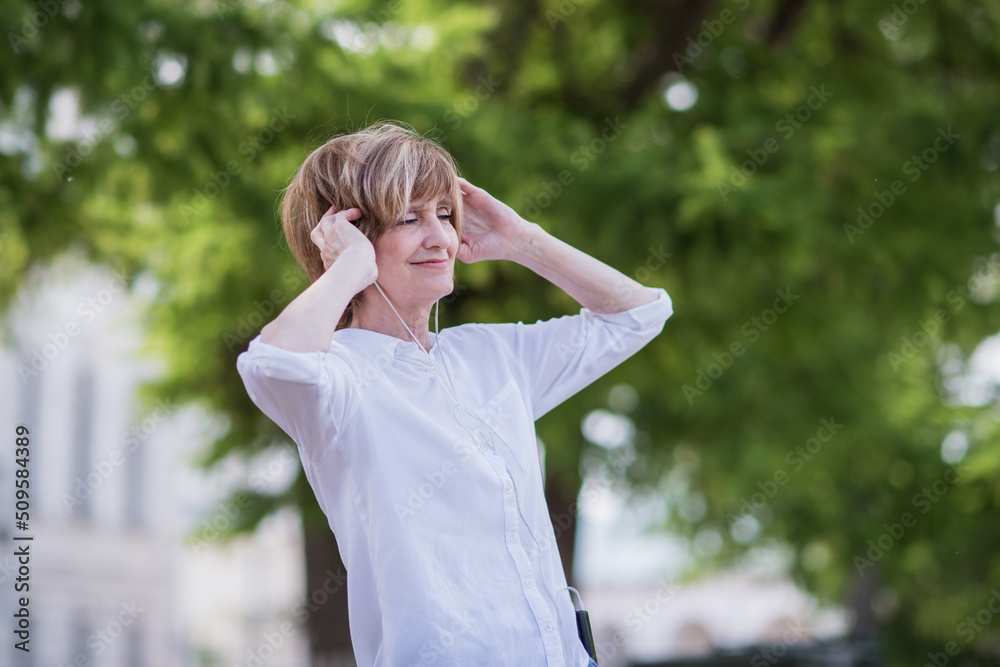 Bottom view elderly happy woman 60 years old in jeans and white shirt puts on headphones, listens to music, dances in a green park with her hands raised up outdoors with green trees in the background Bottom view elderly happy woman 60 years old in jeans and white shirt puts on headphones, listens to music, dances in a green park with her hands raised up outdoors with green trees in the background