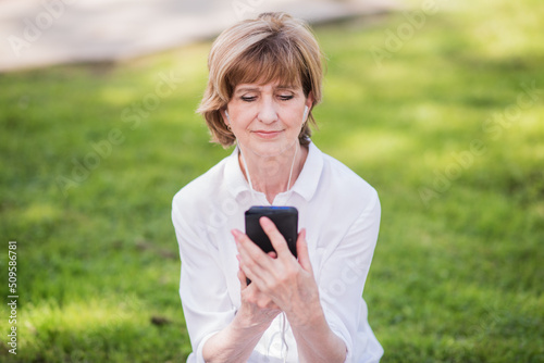 Portrait of a happy elderly woman in a white shirt enjoying music on a mobile phone through headphones in a city park. woman 60 years old listens to music, and has fun in the park outdoors