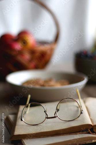 Bowl of strawberries and blueberries, open books with reading glasses, plate of chocolate chip cookies and basket of apples one the table. Selective focus.