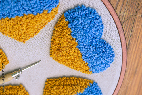 Close up of punch needle embroidered heart in blue and yellow colors and a blurred punch needle on the side. © Uliana