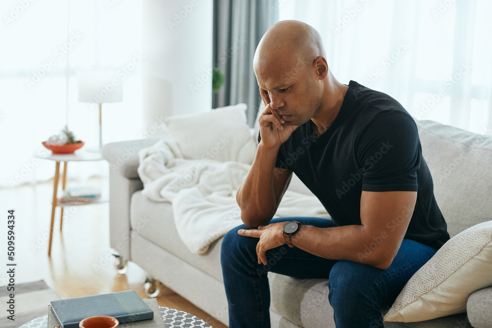 Young worried African American man thinks of his problems while sitting on sofa at home. Young worried African American man thinks of his problems while sitting on sofa at home.