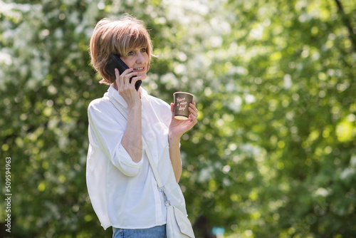 happy senior woman in white shirt looking at phone screen while holding coffee in other hand  walking in city park with coffee in hand 