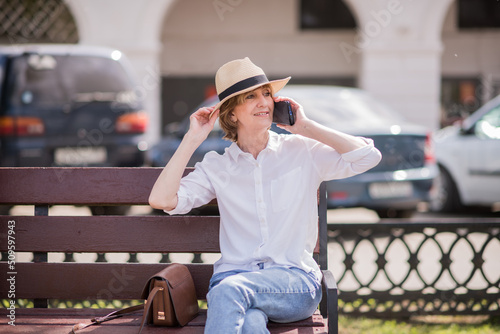 Smiling elderly woman in a wicker hat resting on a bench in a proud park, talking on a mobile phone, rest of an elderly woman 60 years old in her spare time.