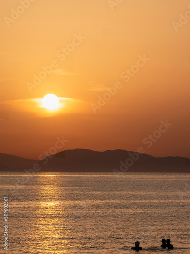Three friends near the sea beach. Panoramic beach landscape and a distant island in the background. Orange and golden sunset sky, calmness, tranquil, relaxing sunlight, summer mood. © Dimitrios