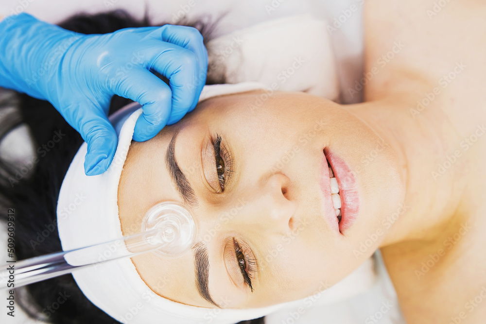 Naklejka premium Closeup photo of professional beautician giving darsonval active electric facial procedure to young woman in beauty salon
