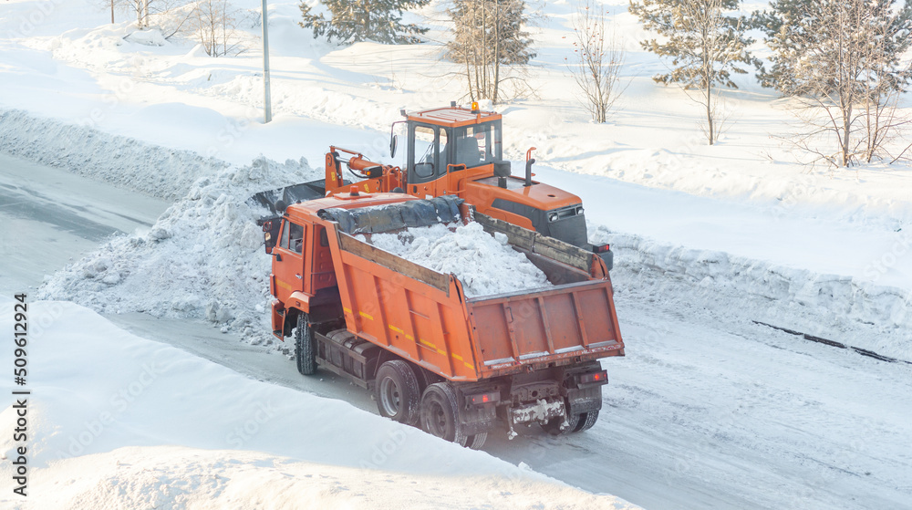 Big orange tractor cleans up snow from the road and loads it into the truck. Cleaning and cleaning of roads in the city from snow in winter. Snow removal after snowfall and blizzards. Big orange tractor cleans up snow from the road and loads it into the truck. Cleaning and cleaning of roads in the city from snow in winter. Snow removal after snowfall and blizzards.