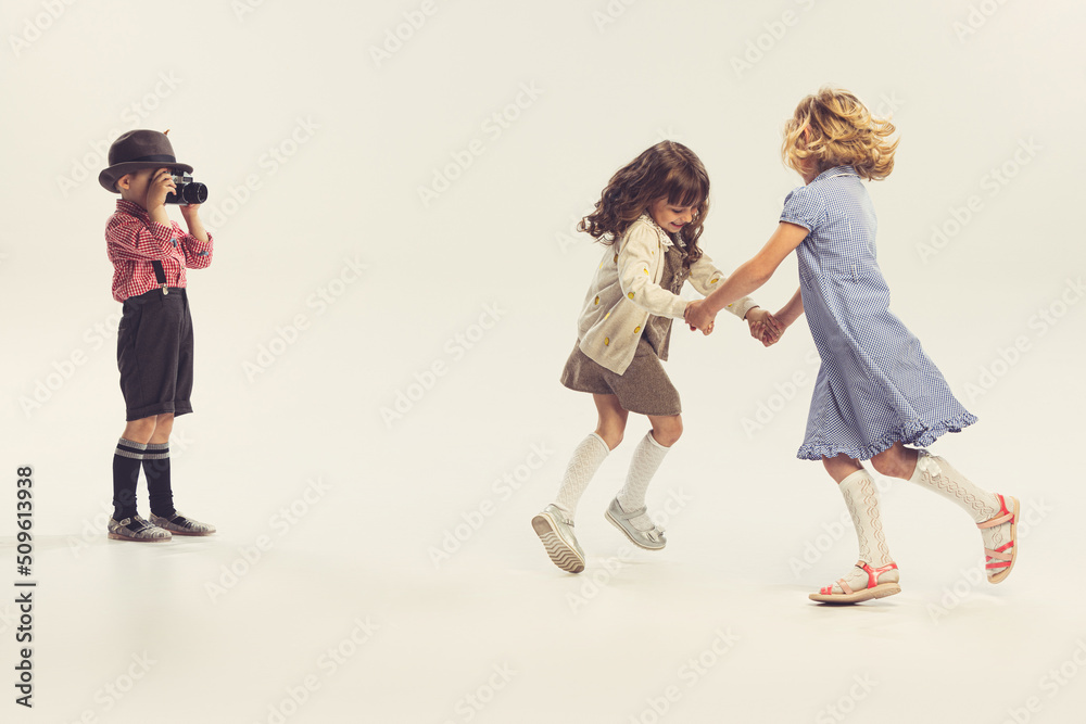 Portrait of three children, little boy taking photo of two cheerful girls holding hands and whirling isolated over grey studio background