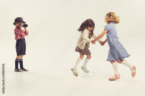 Portrait of three children, little boy taking photo of two cheerful girls holding hands and whirling isolated over grey studio background