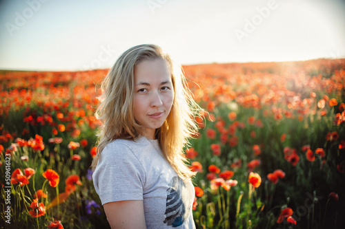 Portrait of a beautiful Caucasian woman smiling and looking into the camera lens during sunset. Outdoor portrait of a smiling white girl. Happy cheerful girl laughs in a field of red flowers