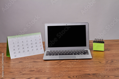 Open laptop with black screen and calendar on wooden desk. Notepad with blank sticky notes.
