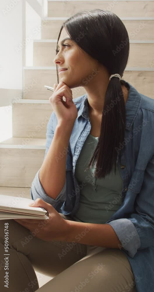 Vertical video of thoughtful biracial woman sitting on stairs and making notes Vertical video of thoughtful biracial woman sitting on stairs and making notes