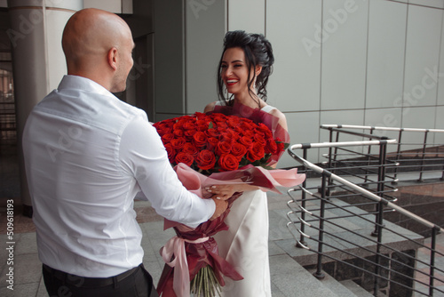 groom in white shirt present with a luxury huge bouquet of red roses to the young happy bride on the gray street building background. wedding celebration concept, free space