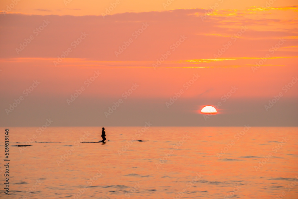 A person walking through calm ocean towards the orange sky and sunset.