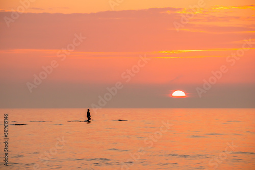 A person walking through calm ocean towards the orange sky and sunset.