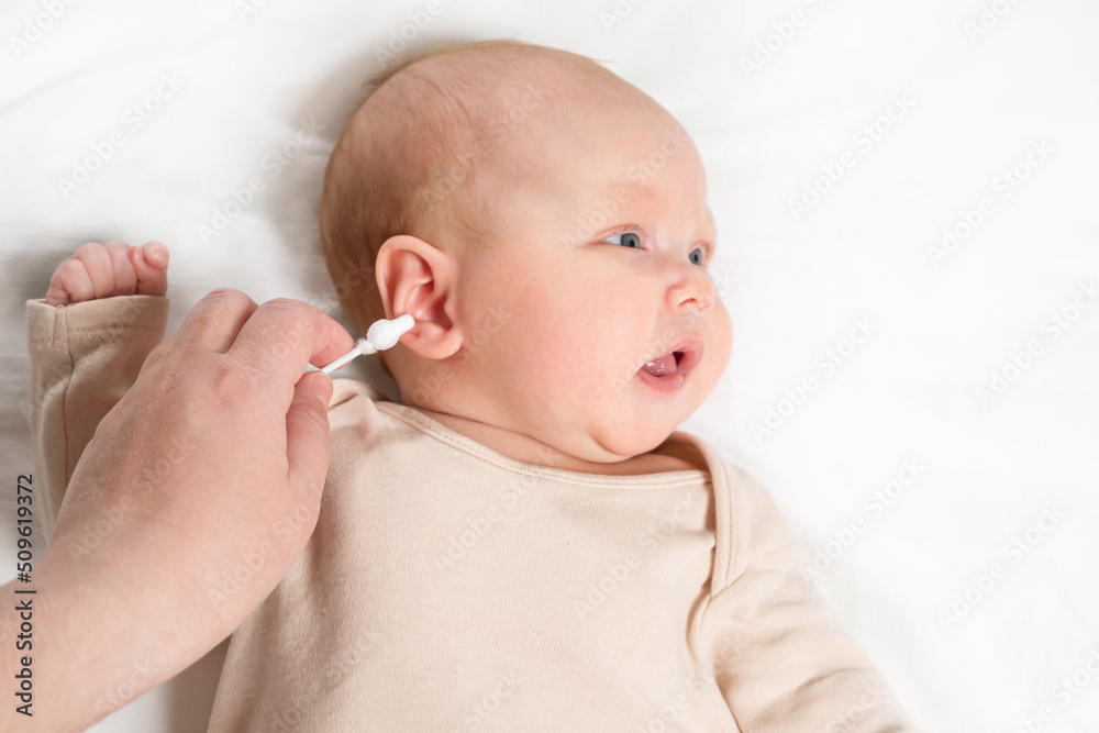Hand of mother cleans ears of baby girl lying on white sheet with cotton swab. Mom takes care of hygiene for newborn kid in bedroom upper close view Hand of mother cleans ears of baby girl lying on white sheet with cotton swab. Mom takes care of hygiene for newborn kid in bedroom upper close view