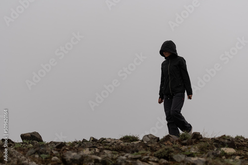 Selective focus shot of a black hooded woman walking alone on rocky terrain in foggy weather.