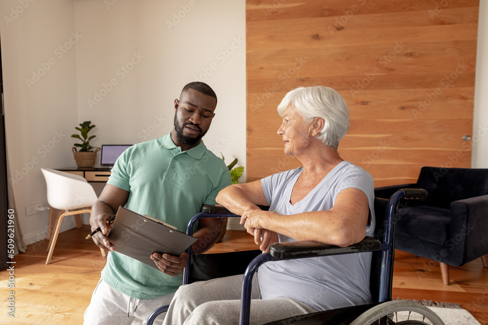Obraz premium African american male physiotherapist with clipboard discussing with senior woman on wheelchair
