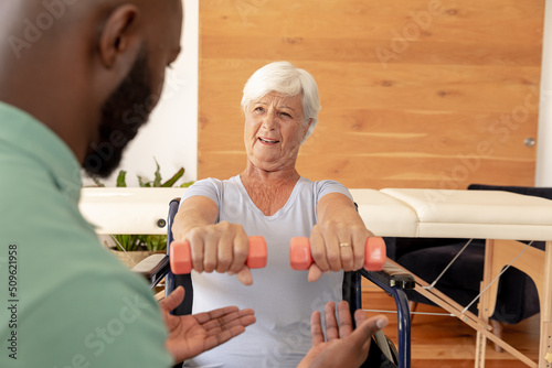 African american male physiotherapist helping caucasian senior woman to do exercise with dumbbells