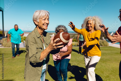 Asian senior woman playing rugby with multiracial friends in yard against clear sky at nursing home