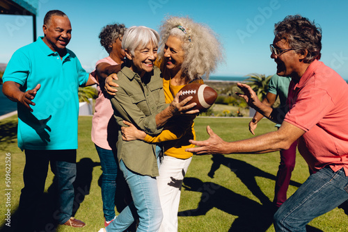 Cheerful multiracial senior friends playing rugby in yard against clear blue sky on sunny day