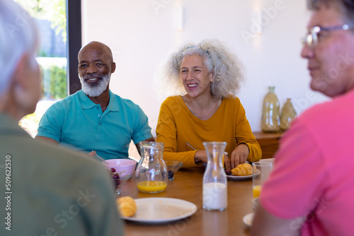 Happy multiracial senior friends talking while having breakfast at dining table in retirement home