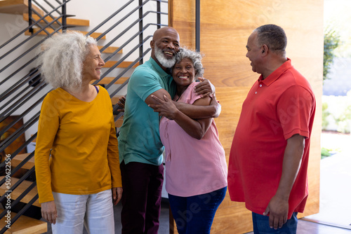 Happy multiracial seniors welcoming friends while standing at doorway in retirement home