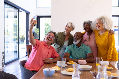 Caucasian senior man taking selfie with multiracial friends while having breakfast at dining table