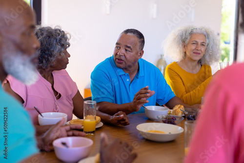 Multiracial senior friends talking and eating healthy breakfast at dining table in nursing home