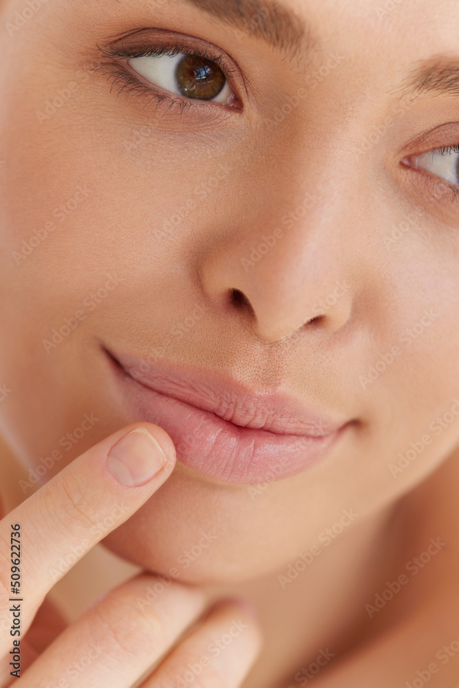 Lips skin care. Beautiful caucasian woman with natural makeup touching with finger her full lips over grey studio background. Concept of treatment, spa and skin care.
