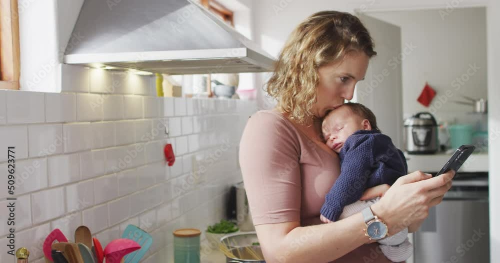 Video of caucasian mother with newborn baby in baby carrier using smartphone in kitchen