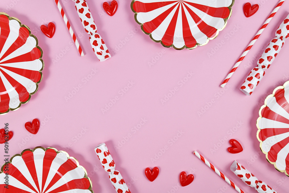 Fototapeta premium Party border with red paper plates, heart ornaments, candy and drinking straws on pink background with copy space