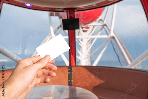 Fototapeta Naklejka Na Ścianę i Meble -  Hand holding white card as entrance ticket or discount with ferris wheel cabin at the background in amusement luna park