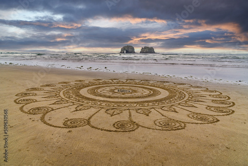 Intricate mandala star sand design on the ocean beach with Twin Rocks in the ocean and beautiful sky.