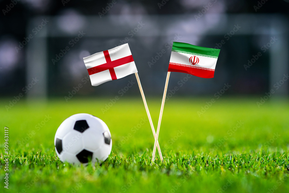 Fototapeta premium England vs. Iran, Khalifa Stadium, Football match wallpaper, Handmade national flags and soccer ball on green grass. Football stadium in background. Black edit space.