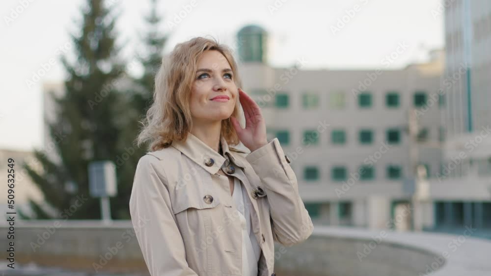 A beautiful slender blonde in a light raincoat stands in the center of the metropolis and enjoys the warm spring weather. Slow motion.