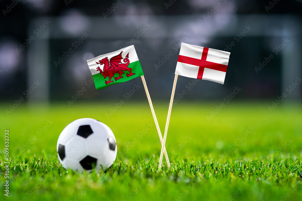 Fototapeta premium Wales vs. England, Ahmad Bin Ali, Football match wallpaper, Handmade national flags and soccer ball on green grass. Football stadium in background. Black edit space.