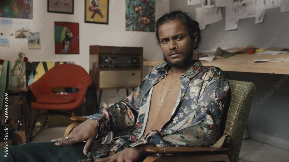 Portrait of handsome young Indian designer or artist in unbuttoned floral printed silk shirt with painted black nails sitting in armchair in creative messy workshop looking at camera Portrait of handsome young Indian designer or artist in unbuttoned floral printed silk shirt with painted black nails sitting in armchair in creative messy workshop looking at camera