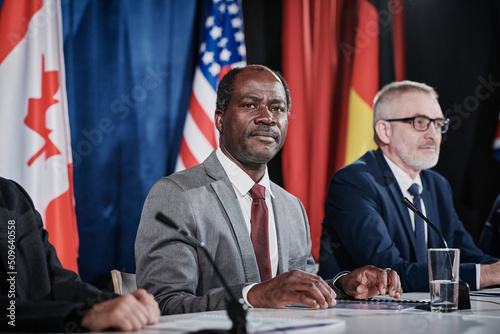 Portrait of African American representative looking at camera while sitting at table in front of microphone with his colleagues during summit