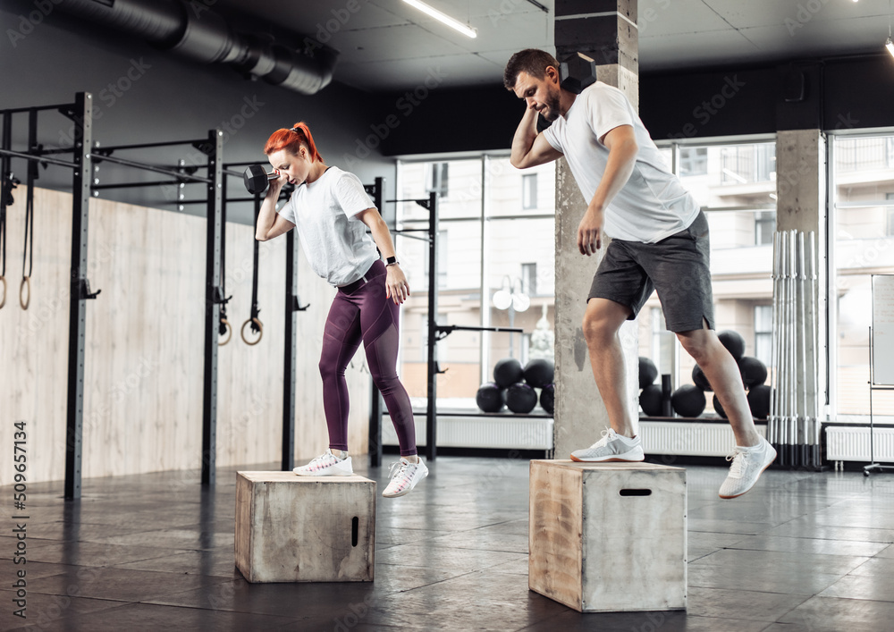 Obraz premium Athletic man and woman practicing lunges together with dumbbell on box in modern gym