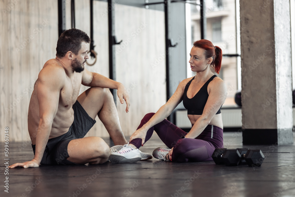 Obraz premium Athletic couple talking while sitting on the floor in the gym. Interaction, communication, healthy lifestyle, bodybuilding and fitness
