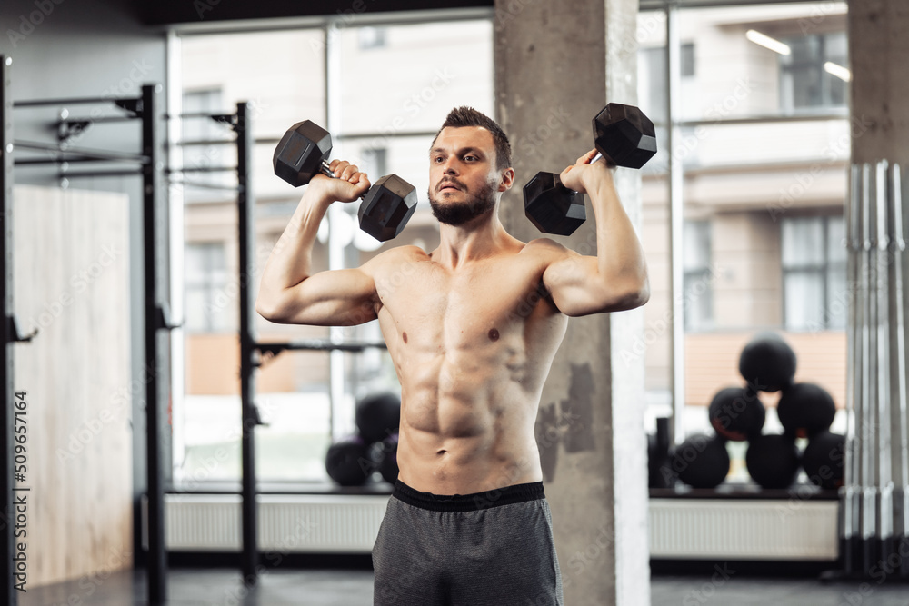 Athletic man with a naked torso trains his shoulders with dumbbells in his hands. Workout in the gym. Bodybuilding and fitness, healthy lifestyle Athletic man with a naked torso trains his shoulders with dumbbells in his hands. Workout in the gym. Bodybuilding and fitness, healthy lifestyle