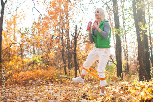 Fototapeta Naklejka Na Ścianę i Meble -  Active and energetic fit blond woman is training with dumbbells and fitness band in the autumn forest. Healthy lifestyle. Outdoor workout