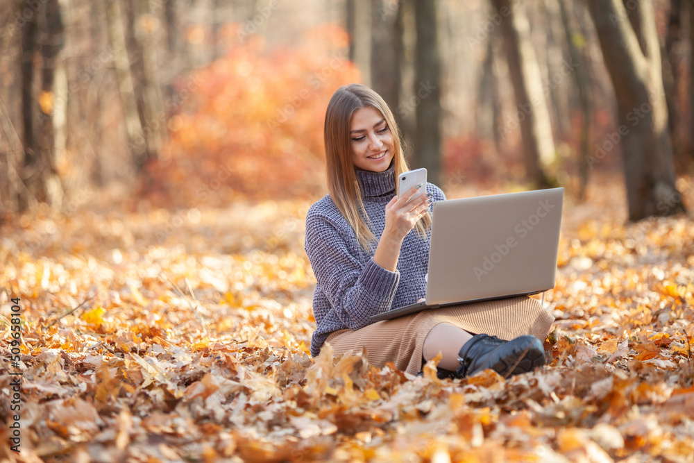 Young female freelancer or student with a laptop and smartphone sits on fallen leaves in autumn forest. Remote work or education Young female freelancer or student with a laptop and smartphone sits on fallen leaves in autumn forest. Remote work or education