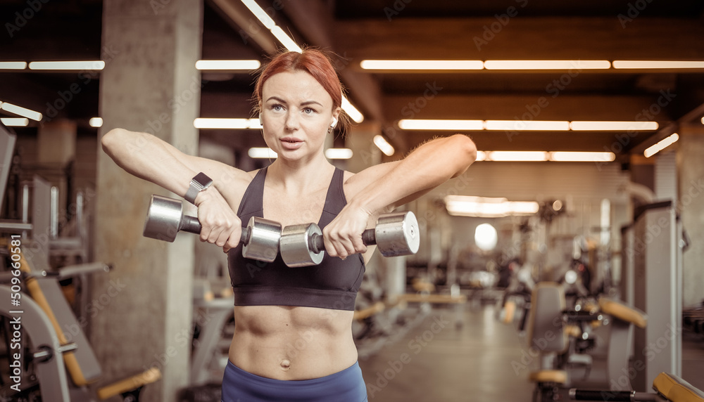 Attractive red-haired fitness woman trains shoulder muscles with dumbbells in a modern gym. Bodybuilding and Fitness Attractive red-haired fitness woman trains shoulder muscles with dumbbells in a modern gym. Bodybuilding and Fitness