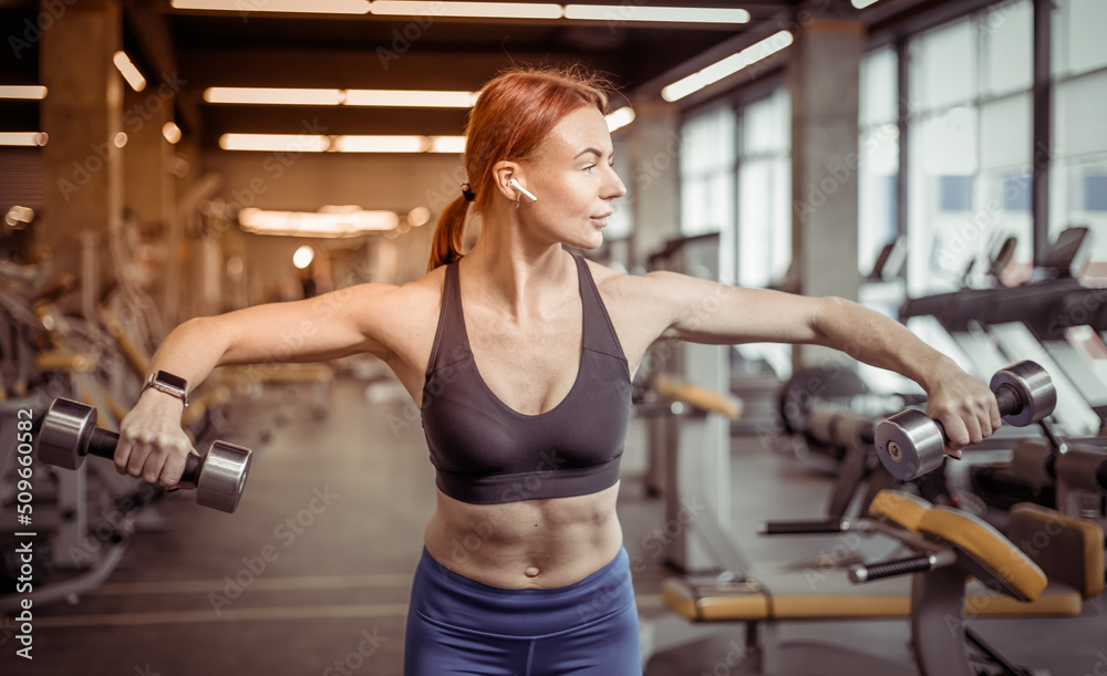 Attractive red-haired fitness woman trains shoulder muscles with dumbbells in a modern gym. Bodybuilding and Fitness Attractive red-haired fitness woman trains shoulder muscles with dumbbells in a modern gym. Bodybuilding and Fitness