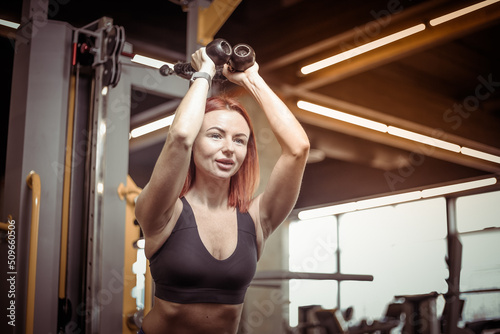 Red-haired young fitness woman in sportswear trains triceps muscles in a cable crossover exercise machine, doing extension of arms from behind head in modern gym. Fitness and bodybuilding concept