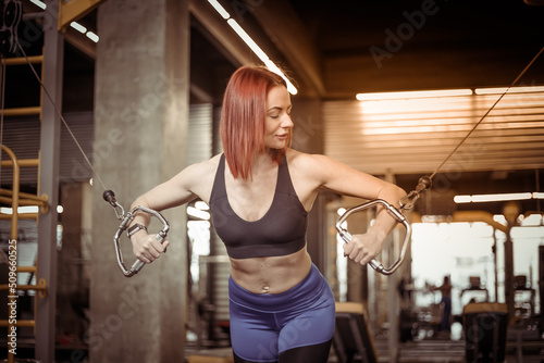 Attractive red-haired fitness woman trains pectoral muscles in a cable crossover exercise machine in a modern gym.