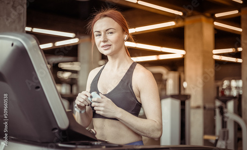 Portrait of a red-haired fitness woman with earphones in a modern gym. Fit slim woman works out on a treadmill, cardio workout, healthy lifestyle.